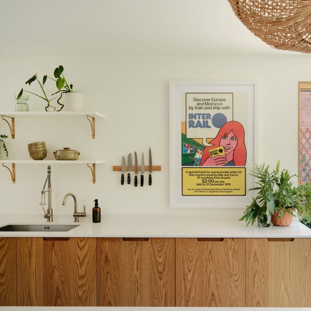 Wide shot of a modern, contemporary kitchen. White walls with accents of green plants in terracotta pots. There is a set of oak-veneered cabinets, with integrated scoop handles. A Caesarstone Calacatta Nuvo polished worktop sits on top of the cabinets. It's a warm and inviting space - designed for the family.